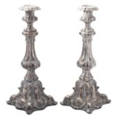 A pair of silvered metal candlesticks, decorated with husks & scrolls, on shaped square bases,