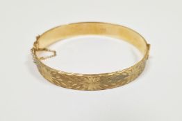 9ct gold hinged bangle with safety chain, one half engraved and the other half plain, 6cm wide,