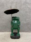 A contemporary carved and painted frog figure holding an umbrella on a carved base. H.92cm