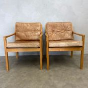 A pair of mid-century beech framed armchairs with distressed tan leather upholstered squab cushions.