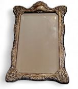 A silver shaped rectangular easel mirror, embossed with foliate scrolls, 37cm high, 24cm wide,