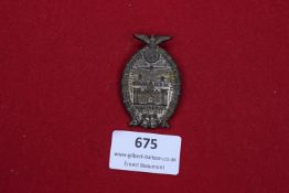 An oval zinc badge with oak-leaf border, eagle and swastika above a castle motif. Inscribed “Hermann