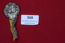 A decorative pressed metal badge with ornate scrollwork border, featuring twin shields at the