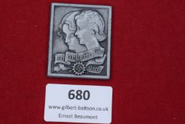 A rectangular zinc badge featuring male and female profiles above a scroll with the inscription 