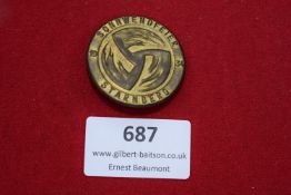 Circular gilt-finish badge, stylised sun-wheel swastika at centre, inscribed 