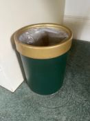A cylindrical wastebasket or bin features a dark green body with a contrasting golden or brass-toned