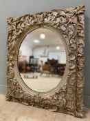 A modern ornate Rococo style wall mirror, oval plate enclosed within a square heavily carved