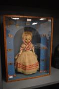 A 19th century wax headed doll, displayed behind an oak glazed cabinet, the background decorated