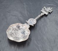 A white metal 'Hanau silver' ornate tea strainer, having scroll work village scene and pierced