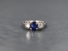 A white metal sapphire and diamond set trilogy ring, having central emerald cut blue sapphire