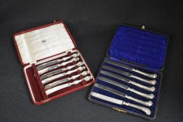 Two cased sets of six silver handled butter knives, both having ornate decoration to the handles,