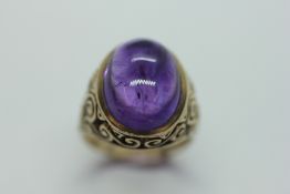 A 9CT GOLD AMETHYST RING, THE OVAL CABOCHON IN A RUB OVER SETTING, THE SHOULDERS OF SCROLLED DESIGN.
