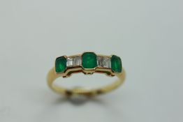 AN UNMARKED YELLOW METAL RING SET WITH THREE CUSHION CUT EMERALDS ALTERNATING BETWEEN FOUR SMALL