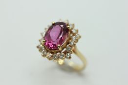 A 14CT YELLOW GOLD DRESS RING SET WITH CENTRAL OVAL PINK STONE SURROUNDED BY TWENTY SMALL DIAMONDS.