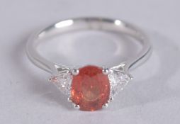 A PLATINUM TRILOGY RING SET WITH A CENTRAL OVAL ORANGE SAPPHIRE FLANKED BY BRILLIANT CUT DIAMONDS.
