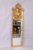 A DECORATIVE GILT FRAMED NARROW MIRROR, the upper section with a bust of a young woman.