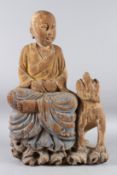A CARVED WOOD BUDDHIST FIGURE OF PATACARA sitting cross legged, a lion dog by his side. 60cms high.