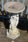A WEATHERED COMPOSITE BIRD BATH, raised on a support with three scantily clad ladies, on an