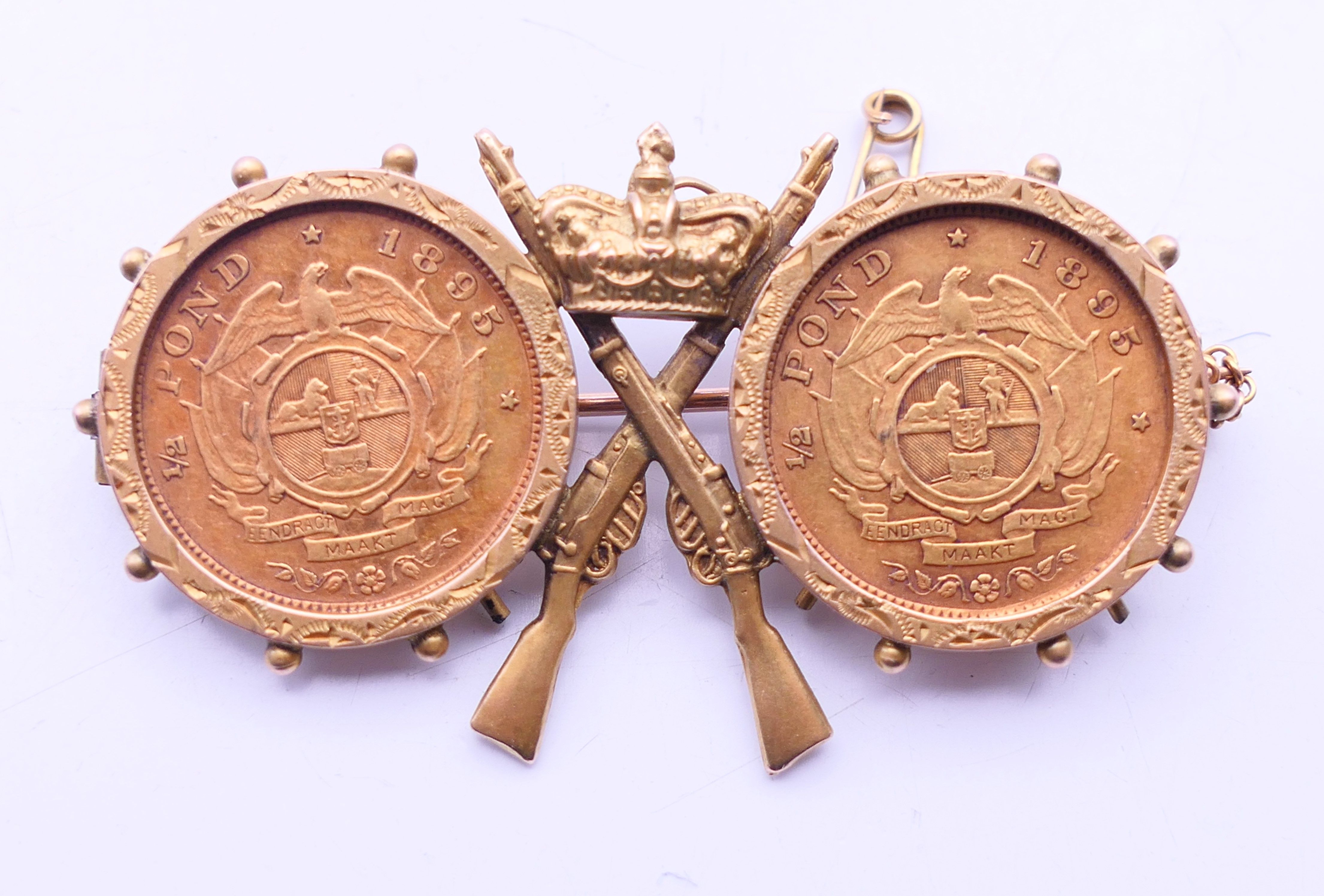 A 9 ct gold military brooch set with two 1895 South African half-pond coins. 5 cm wide x 3 cm high.