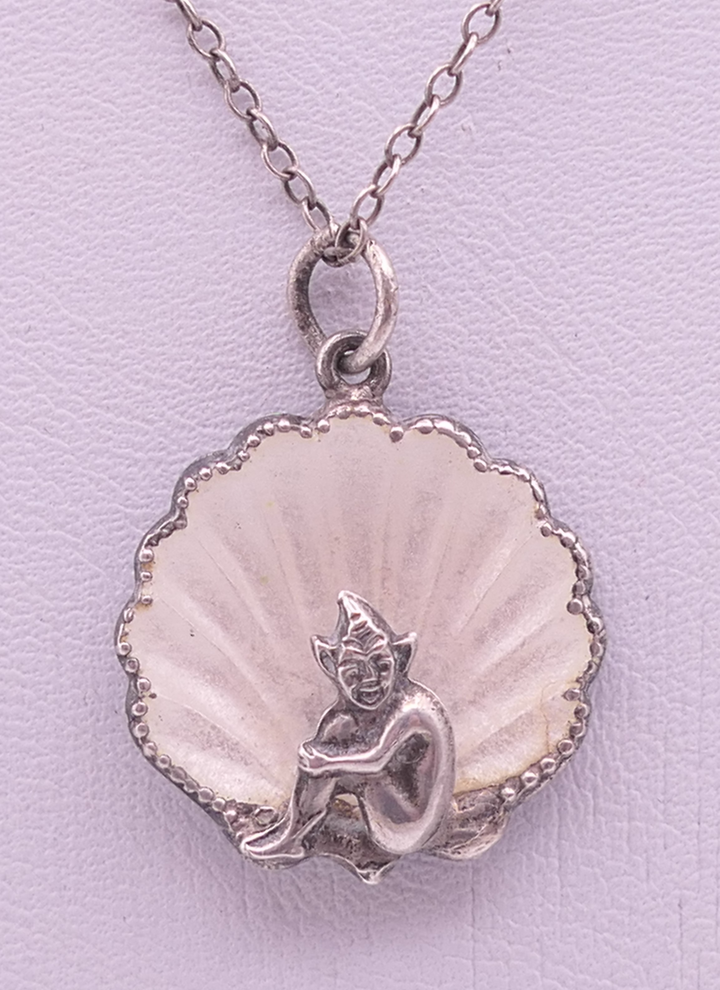 A silver and enamel pixie in a shell pendant on a silver chain. Pendant 2 cm high, chain 46 cm long.