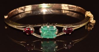 A yellow metal bangle set with an emerald cut emerald of approximately 4.8ct and two garnets, 11.8g,