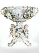 A German porcelain tazza in the manner of Meissen, the twin-handled pierced bowl supported by putti,