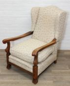 An antique style high back low armchair with carved and exposed oak arms and patterned cream