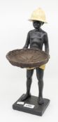 A figure of an African boy holding a basket with pith helmet on.
