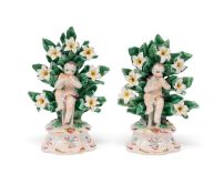Two Bow miniature figures of putti, c.1765, each seated before flowering bocage with one knee raised