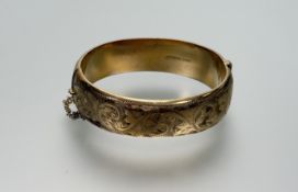 A 9ct gold metal core engraved, stiff hinged bangle with clip fastening and safety chain: d. x 6cm