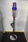 A Dyson v15 detect animal vacuum cleaner on stand with a stair head attachment and plug.
