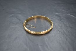A 9ct gold hinged bangle, having decorative scroll engraving to one side, with safety chain,