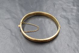 A 9ct gold hinged bangle, having decorative foliate engraving to one side, with safety chain, marked