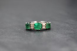 A white metal emerald and diamond set ring, the central cushion cut emerald flanked by a princess