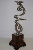 A modern spiral-form brushed metal candlestick rising from a stepped dark-stained wooden plinth, the