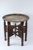 AN EASTERN ENGRAVED BRASS CIRCULAR TRAY, on a carved folding wooden stand. 58cms diameter x 56cms