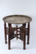 AN EASTERN ENGRAVED BRASS CIRCULAR TRAY, on a carved folding wooden stand. 58cms diameter x 56cms