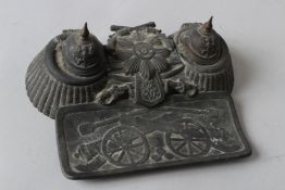 AN ANTIQUE MILITARY THEME DESK INKWELL SET depicting a FIELD CANNON, CROSS SABRES, HELMETS etc. 7ins
