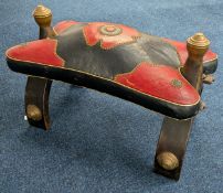 A Moroccan style camel saddle stool, with red and black upholstered seat and brass details.