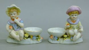A pair of porcelain salts, each surmounted with a child. Each 10 cm high.