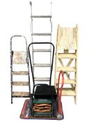 A group of metal and wooden stepladders, push-along garden mower and a flatbed trolley.