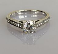 A round brilliant-cut diamond ring with pave-set decoration to shoulders on an 18ct white gold setti