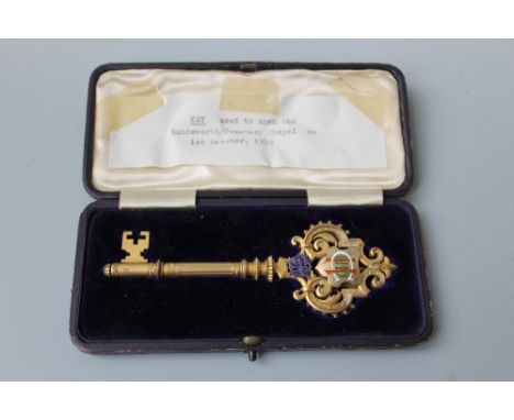 A CASED SILVER GILT AND ENAMEL PRESENTATION KEY, the fancy Gothic style top, with central shield of enamelled intertwined ini