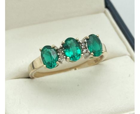 A 9ct gold diamond and created emerald dress ring. 3 oval cut emeralds in a trilogy setting, with 2 small diamonds between ea