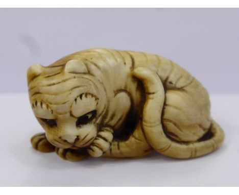 A Meiji Period Japanese Ivory Netsuke, depicting a crouching tiger licking his paw, character marks to the tigers haunches, a