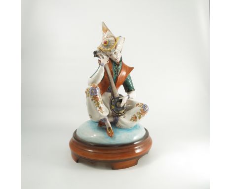 A 1920's Italian porcelain model, of a seated figure, wearing a headdress, playing a stringed instrument, on separate base, m