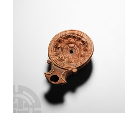1st century A.D. A terracotta oil lamp composed of a circular discus, tapering sidewalls, volute nozzle embellished with scro