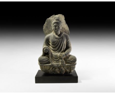 2nd-3rd century AD. A carved schist statue of Buddha sitting cross-legged on a dais with drapery and crouching figures, the b