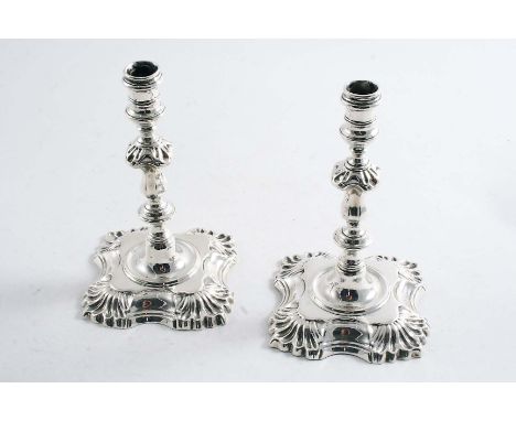A PAIR OF EARLY 20TH CENTURY CAST TAPERSTICKS in the George II style with shaped square bases, shell decoration and spool-sha
