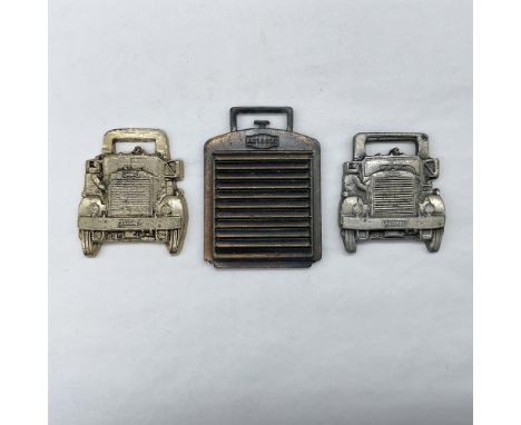 Metal finish with embossed design and lettering. First fob front view: car-shaped in a light golden-color, with Autocar as th